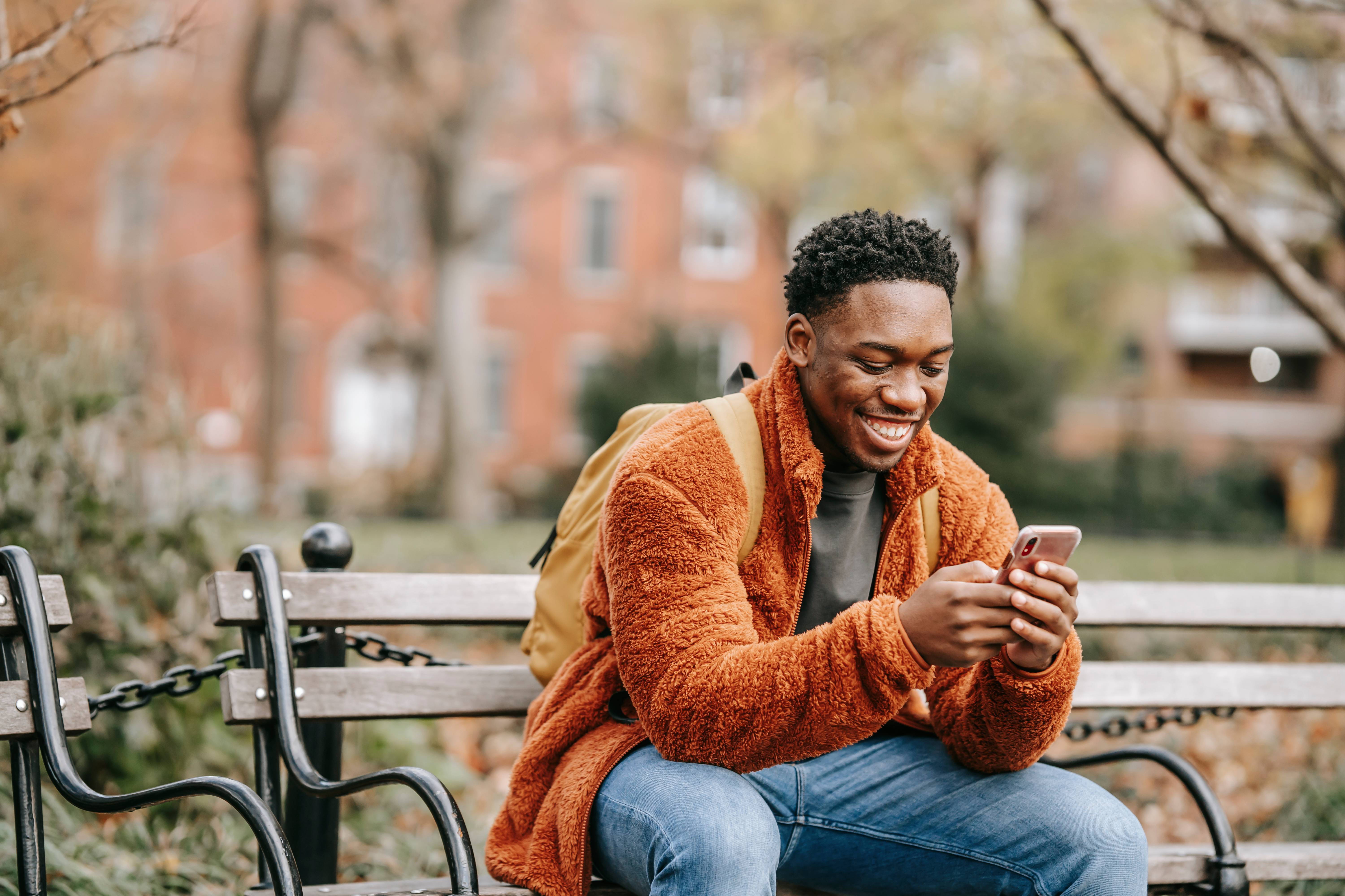 Man with orange jacket sitting on park bench and looking at his phone