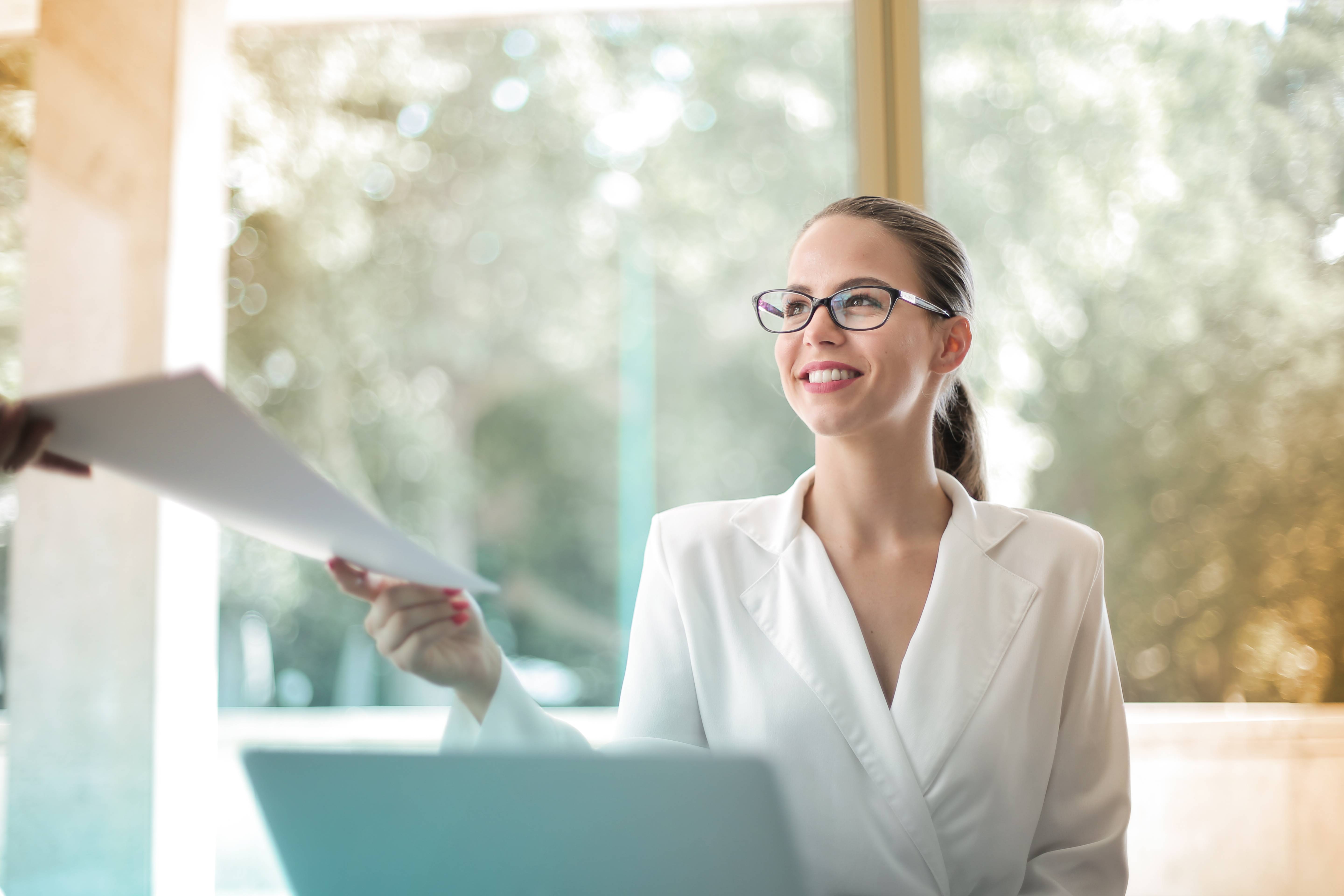 Woman sitting in front of a laptop handing over a document